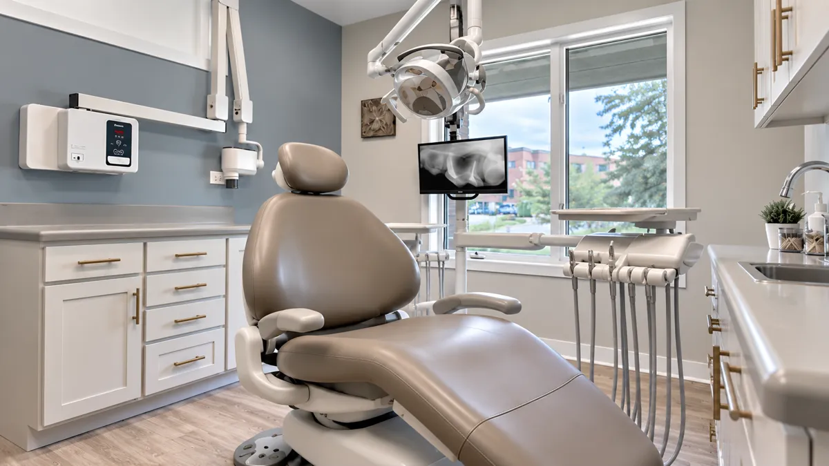 Treatment room with a taupe dental chair, gold cabinet handles and a large window onto trees